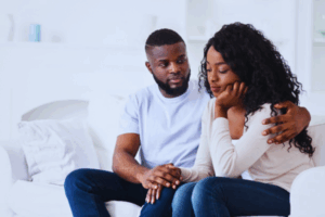 A Nigerian couple sits quietly on a sofa, holding hands in a moment of emotional connection, their expressions reflecting the silent struggle of infertility depression, with soft sunlight streaming through a window in a cozy, warmly lit living room.